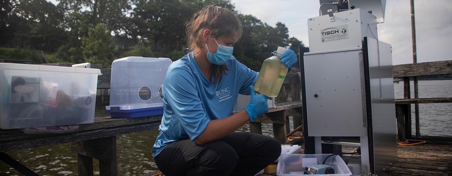 unc marine biologist examines bottle of water with algal blooms while kneeling outside by pier