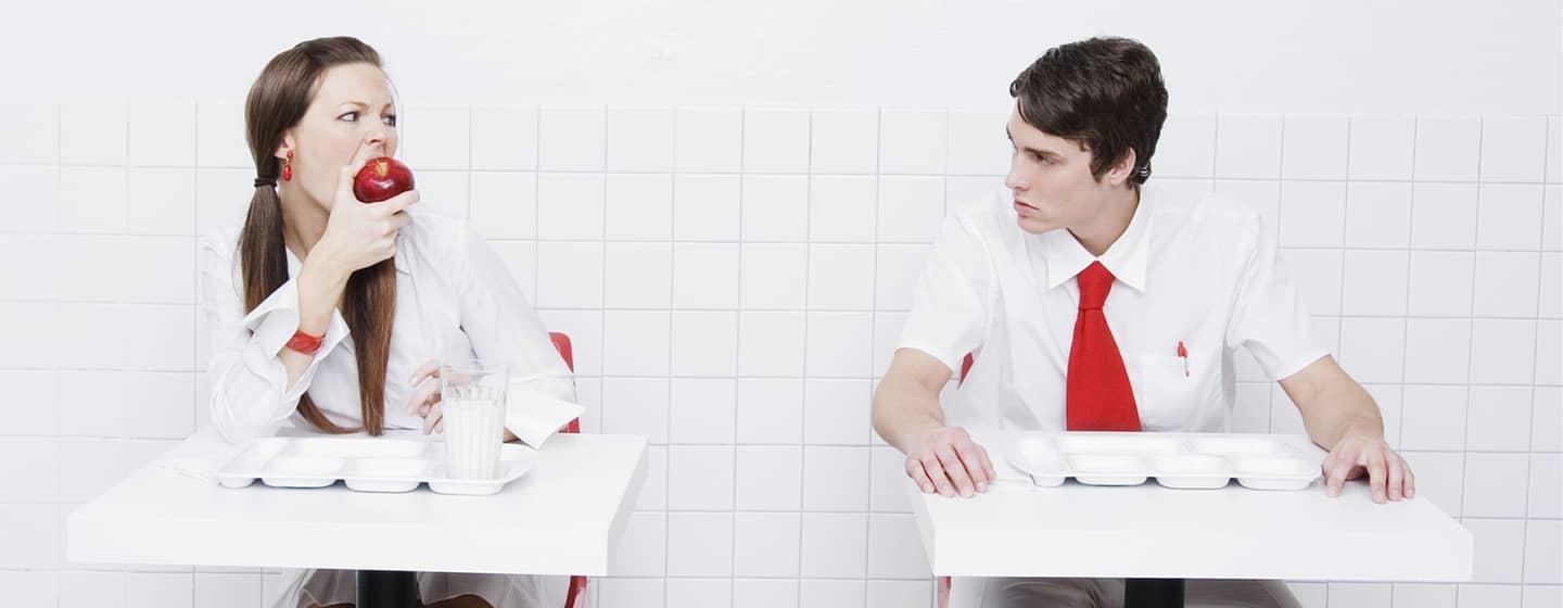 Two people sit at small tables. The woman on the left is biting into an apple while the man on the right looks on with concern.