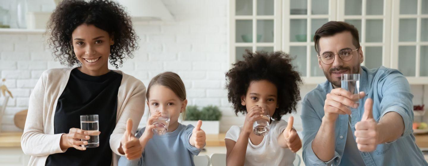 Two parents and their kids drink water at the kitchen table while giving a thumbs up to the camera.