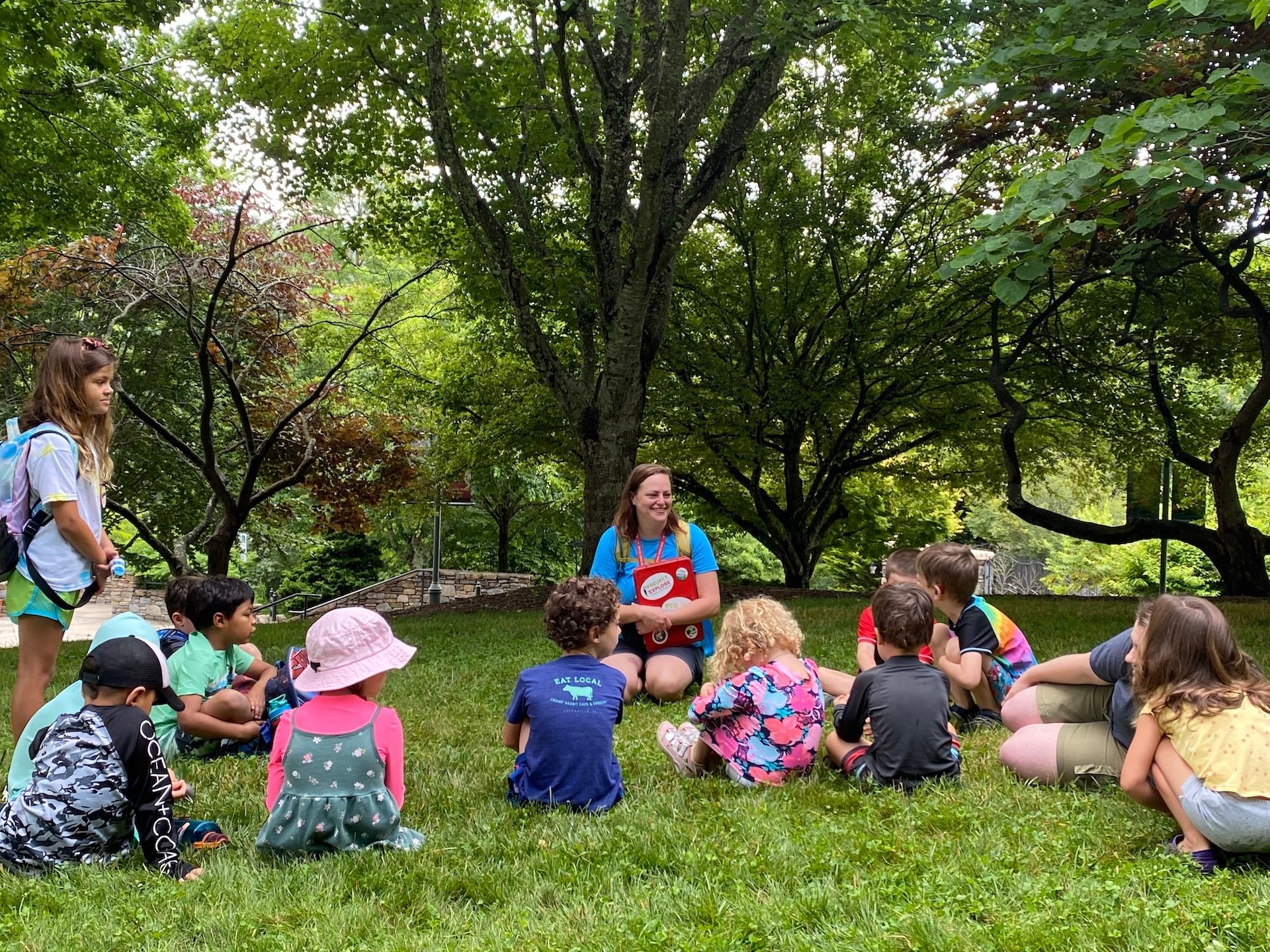 Children sit on the grass around an instructor, outside at the NC Arboretum