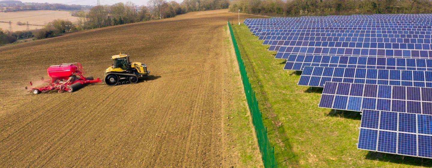 A tractor pulls a seed spreader on the left half of a farm field, and solar panels line the right half of the field.