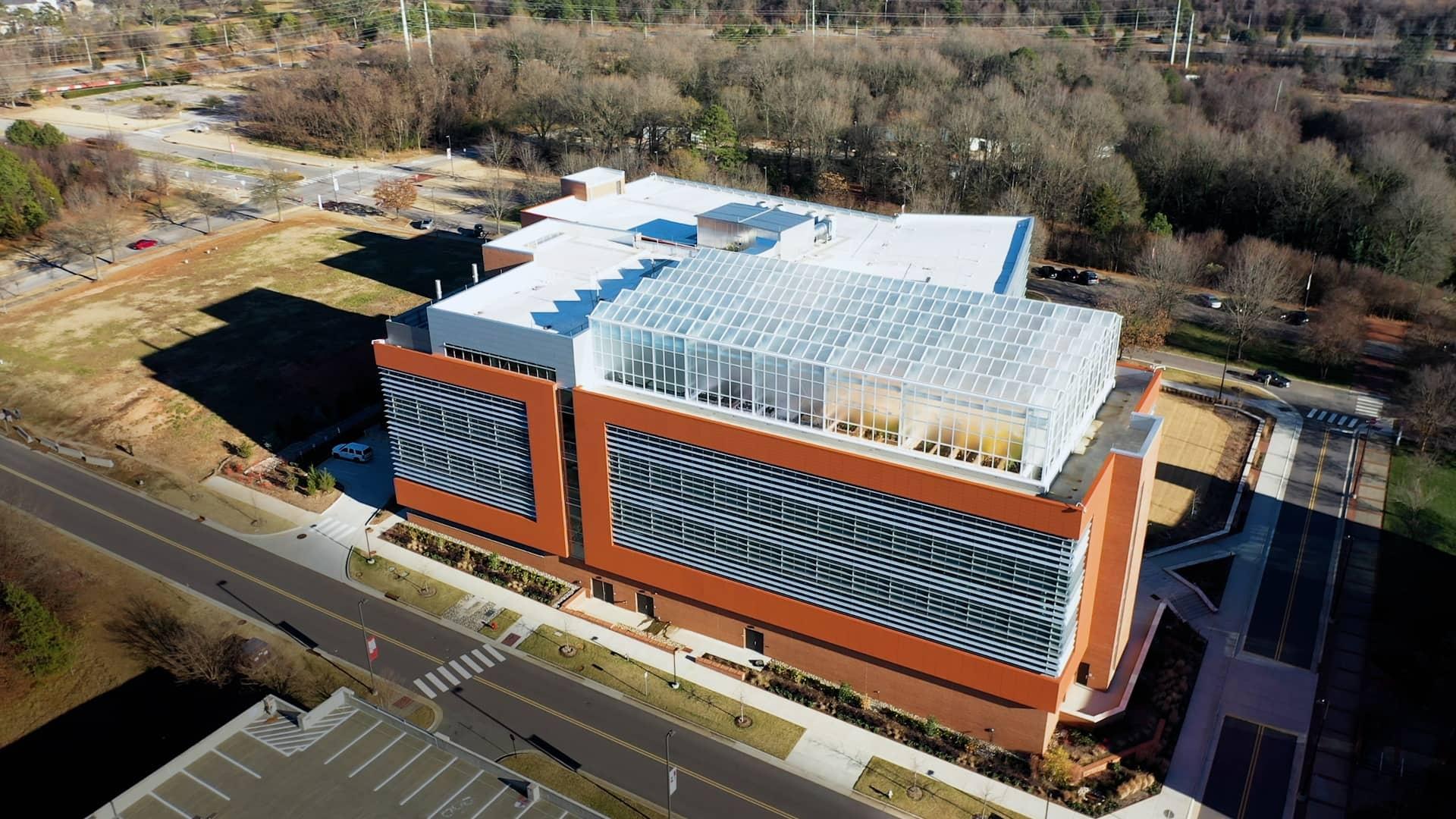 An aerial view of an orange-brick building with sides full of windows and translucent glass greenhouses built on the rooftop.