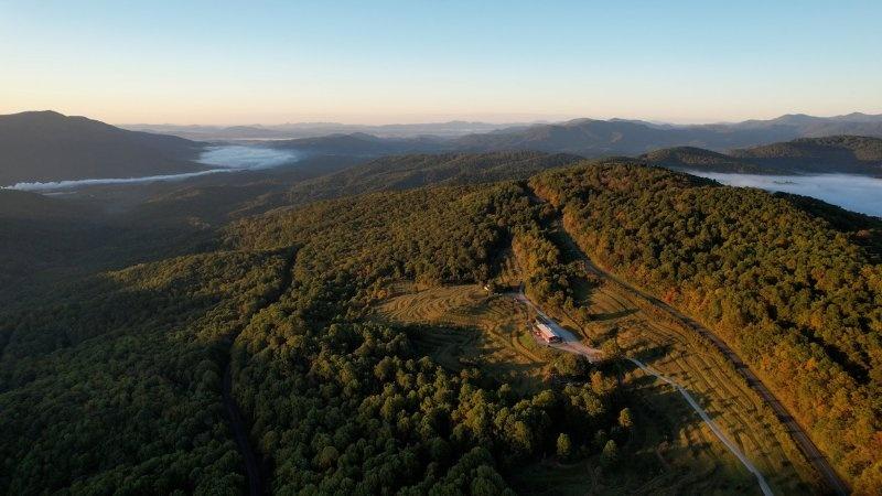 An aerial image at sunrise of tree-covered mountains with mist lingering in between them. The sun is hitting a ridgeline that has a two-lane highway on it. Below the highway are a few buildings and a neatly laid out apple orchard. Farther down from the orchard, railroad tracks cut through the trees leaving a visible gap.
