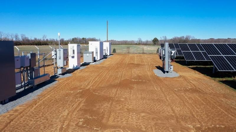 Looking down an aisle between a collection of solar panels (right) and their associated battery-storage infrastructure (left). The earth is a red clay, and green agricultural fields can be seen beyond the fence around the equipment. The batteries are white metal boxes lined up in a row with other gray and white boxes holding electrical equipment.