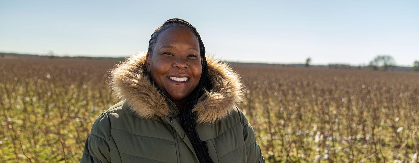 Ajulo Othow, a black woman with cornrows and a green puffer coat, stands in a field while smiling.