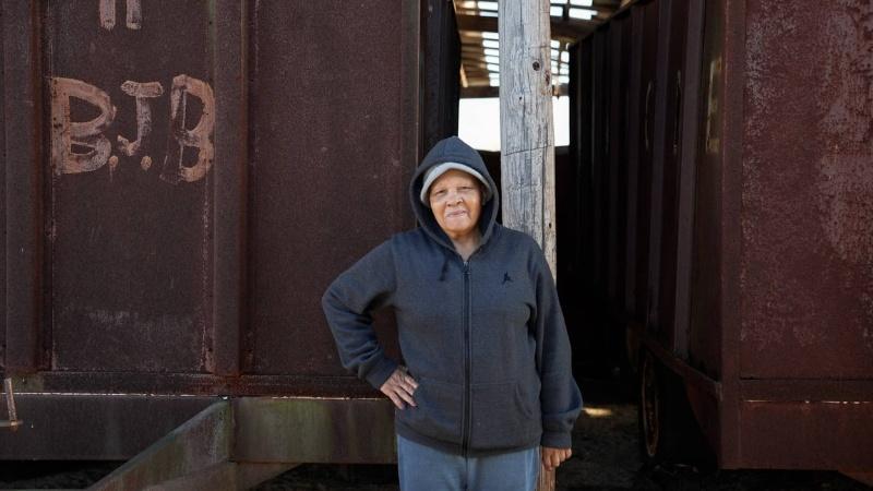 An older Black woman stands in front of two rust-colored industrial containers. She wears a dark gray hoodie with the hood up and a knit hat under it, and has her hand on her hip and a smile on her face. The container in the left of the image has the initials BJB spray painted on it.
