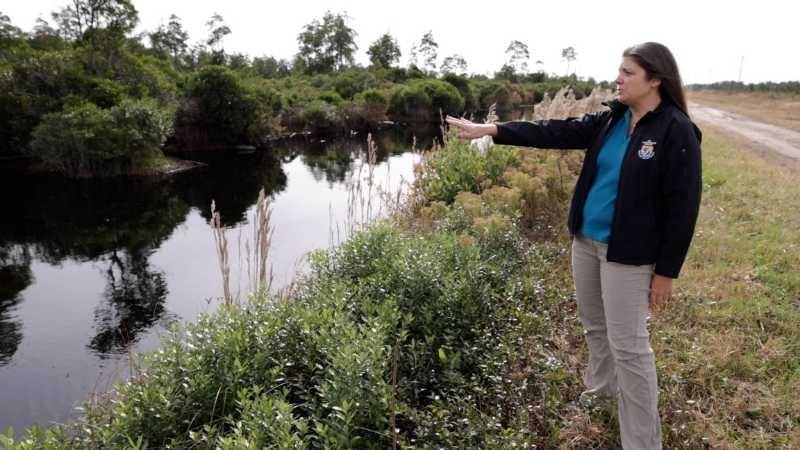 A woman stands next to and gestures at a ditch full of dark black water.