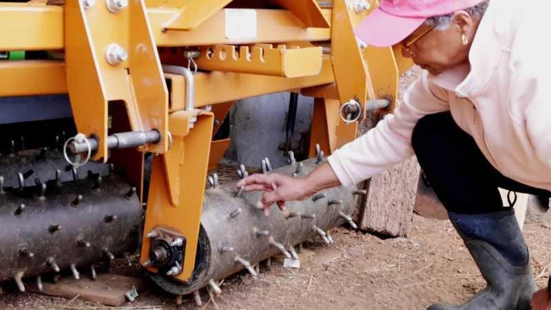 A woman in a pink fleece, pink hat, black pants and rubber work boots kneels in front of an orange metal piece of farming equipment with round metal barrels hanging below it at soil level, that have dull spikes sticking out at even intervals.