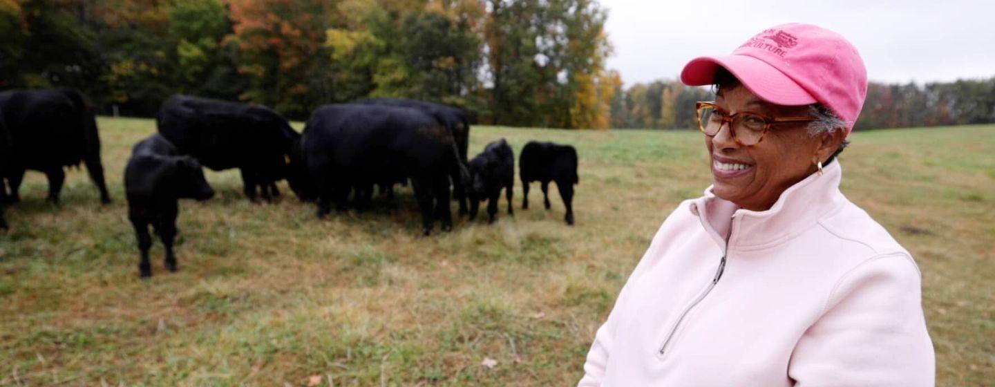 A woman in a light pink zip-up fleece and bright pink hat with a small herd of black cows behind her. They are in a wide open field surrounded by trees with a touch of fall color.