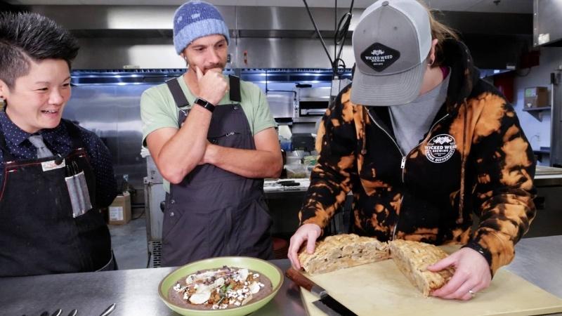 Three people stand around a stainless-steel table in an industrial kitchen. One wears a chef’s apron, one is in overalls and a T-shirt, and the other wears a hooded, tie-dye sweatshirt and a baseball cap. The person with the baseball cap pulls apart two halves of what looks like a latticed apple pie. Next to that on the table is a green bowl with food in it that includes tiny, popped kernels of sorghum, chips, greenery and a purple base. Everyone smiles as they look at the pie.