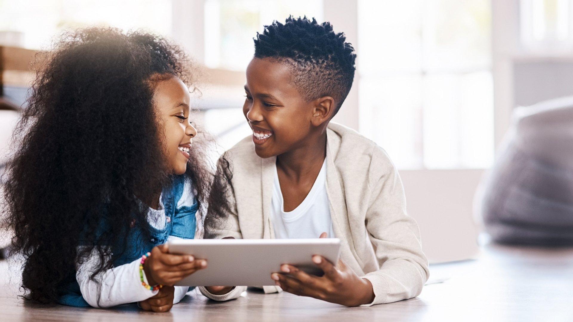 Two Black children facing each other and smiling while playing on a tablet.