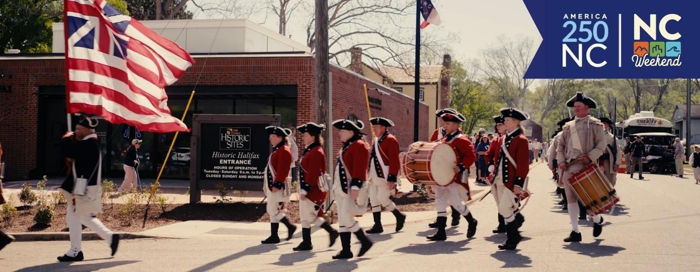 Revolutionary War reenactors marching down a street with the America 250 North Carolina and NC Weekend logo in the top right corner.