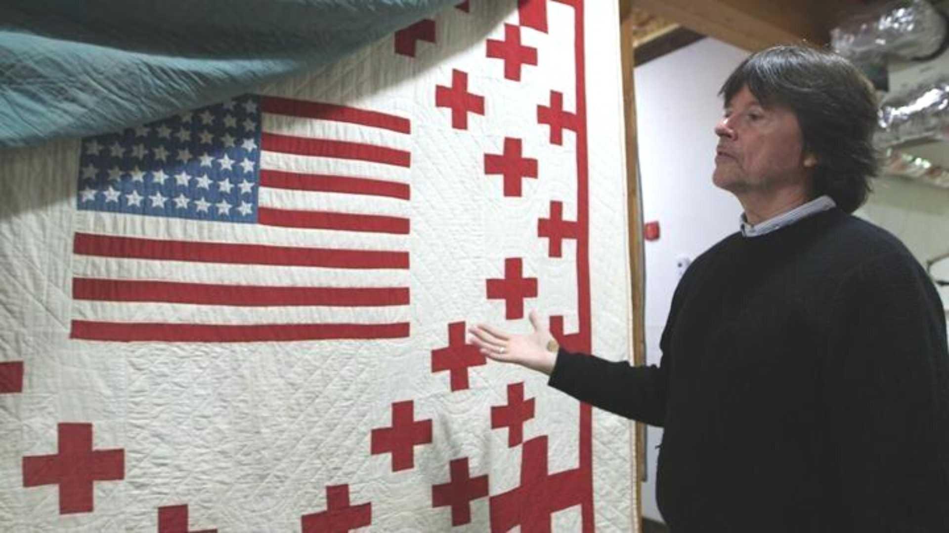Ken Burns stands near and gestures towards a quilt from his collection.