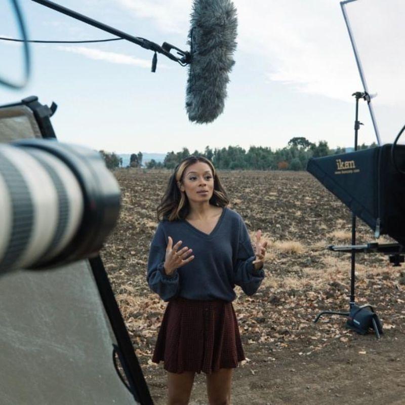 Host of Weathered, Maiya May, stands in front of a field while a boom mic is above her and she's being filmed.