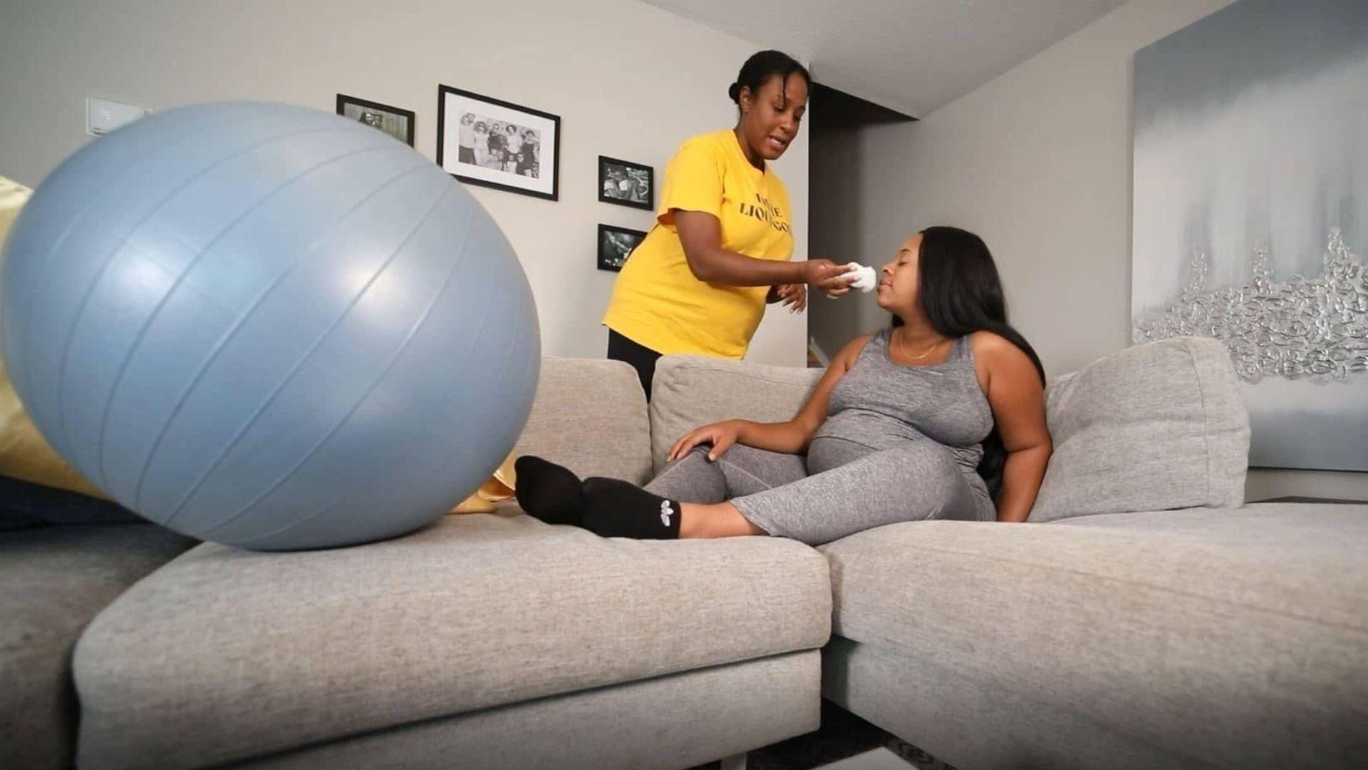 A Black woman on the couch pregnant with a medicine ball next to her. Another black woman helps her with wiping her nose.