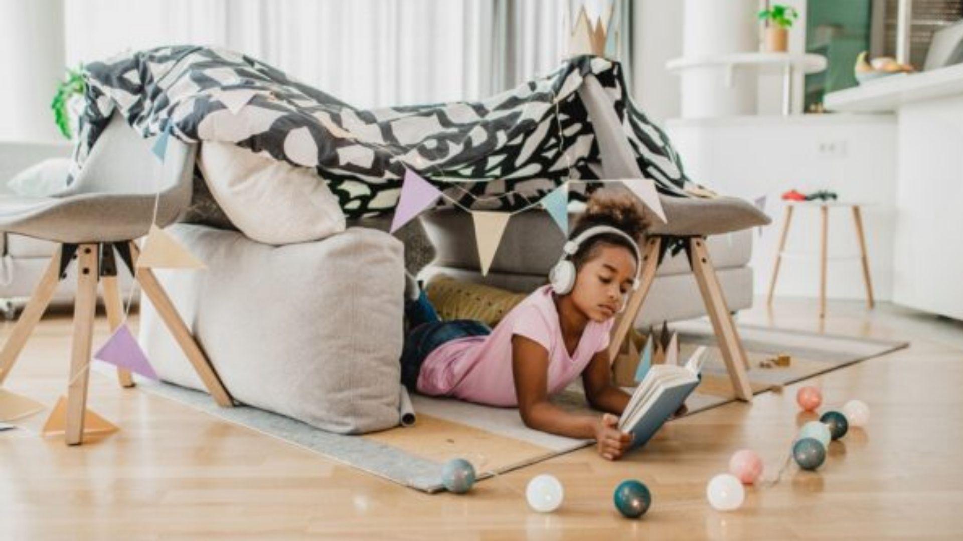 A little girl laying in a pillow fort with headphones on and reading a book.