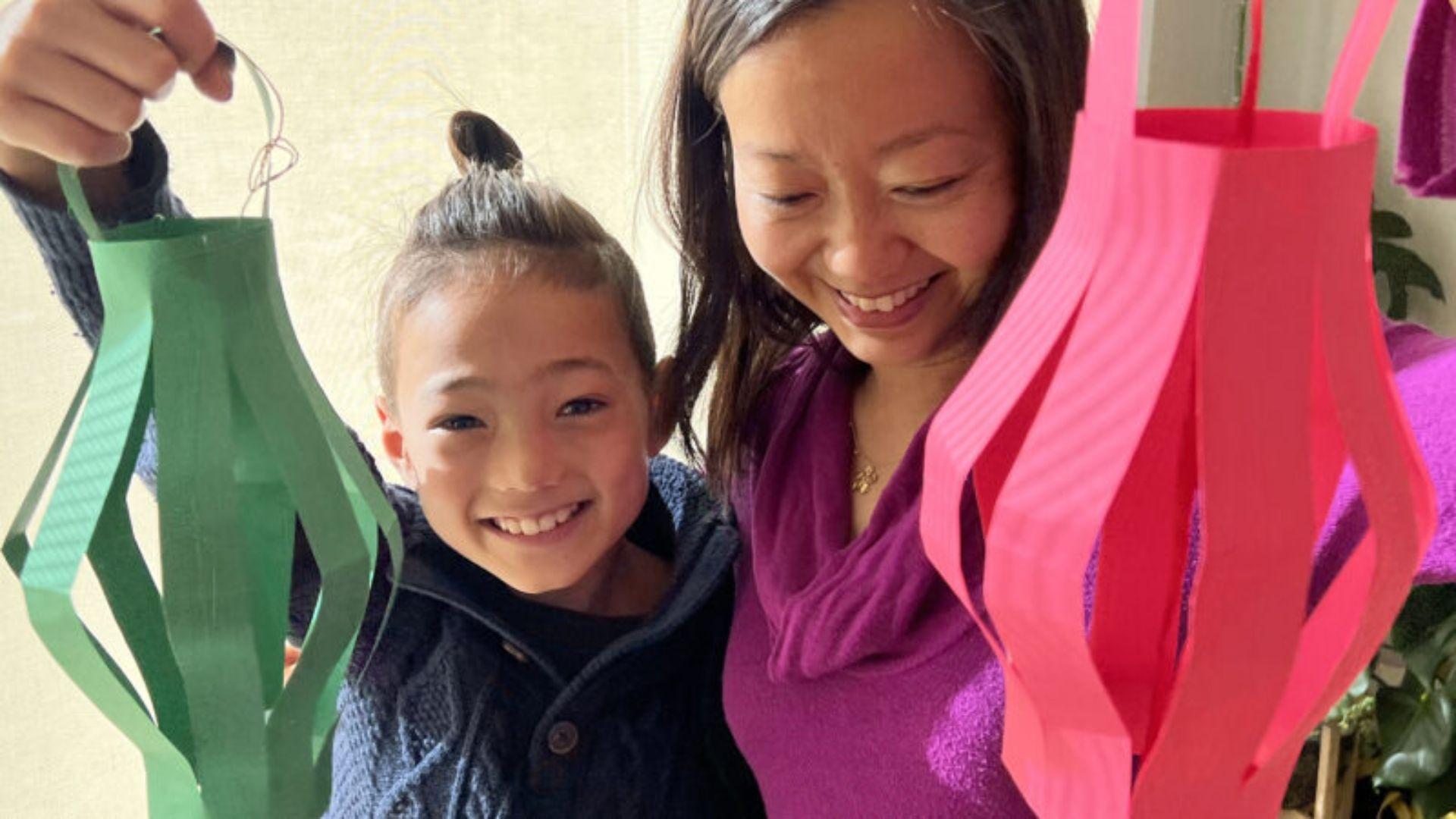 A Child and their parent hold up homemade paper lanterns while smiling.