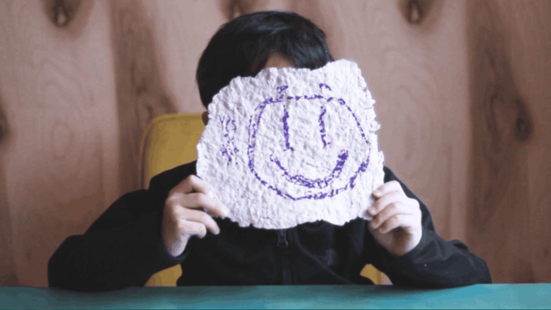 A child with black hair holding up homemade paper to cover their face, with a purple drawn smiley face on it.