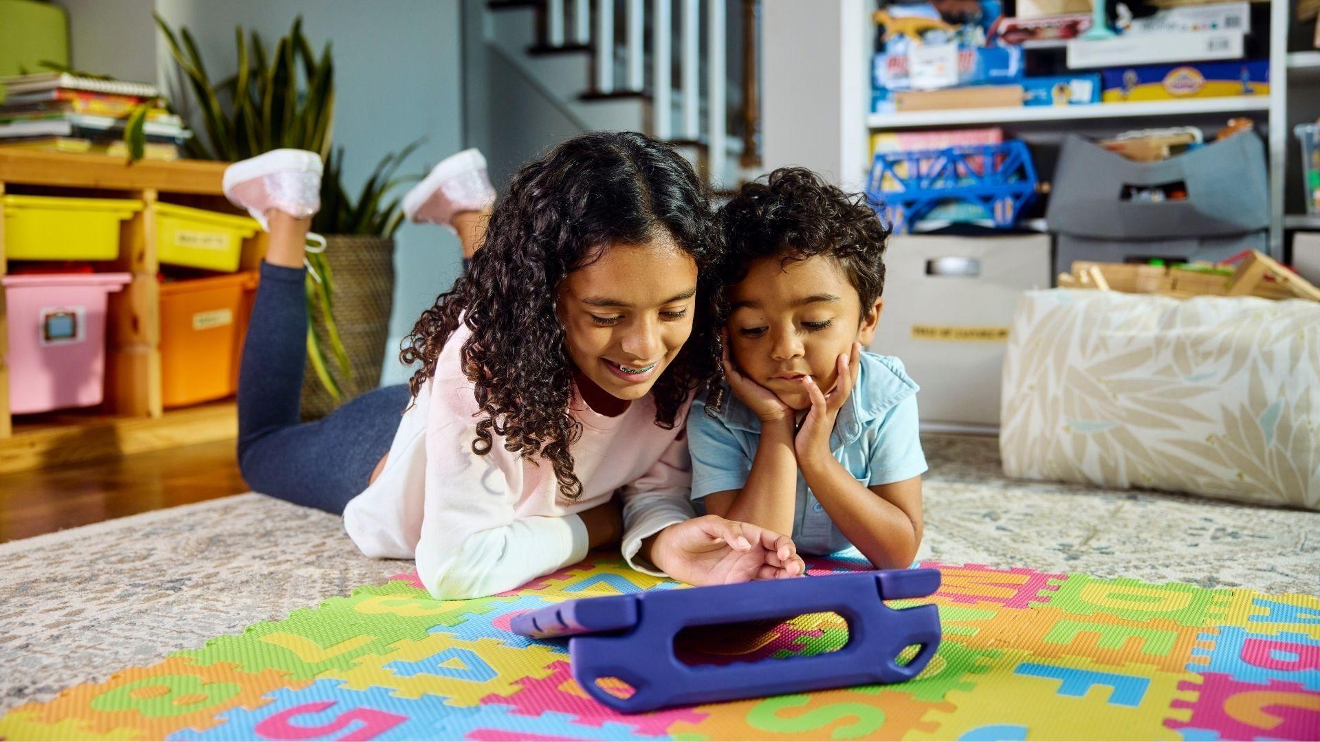 Two children laying on a playroom floor together smiling down at a tablet.