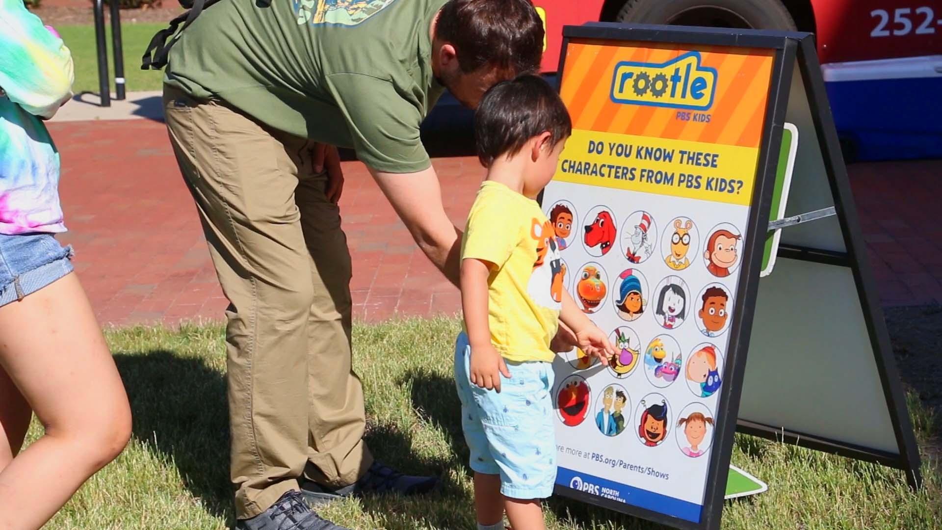 A child points at Rootle PBS KIDS characters on a poster quiz at a Rootle Roadster Tour stop.