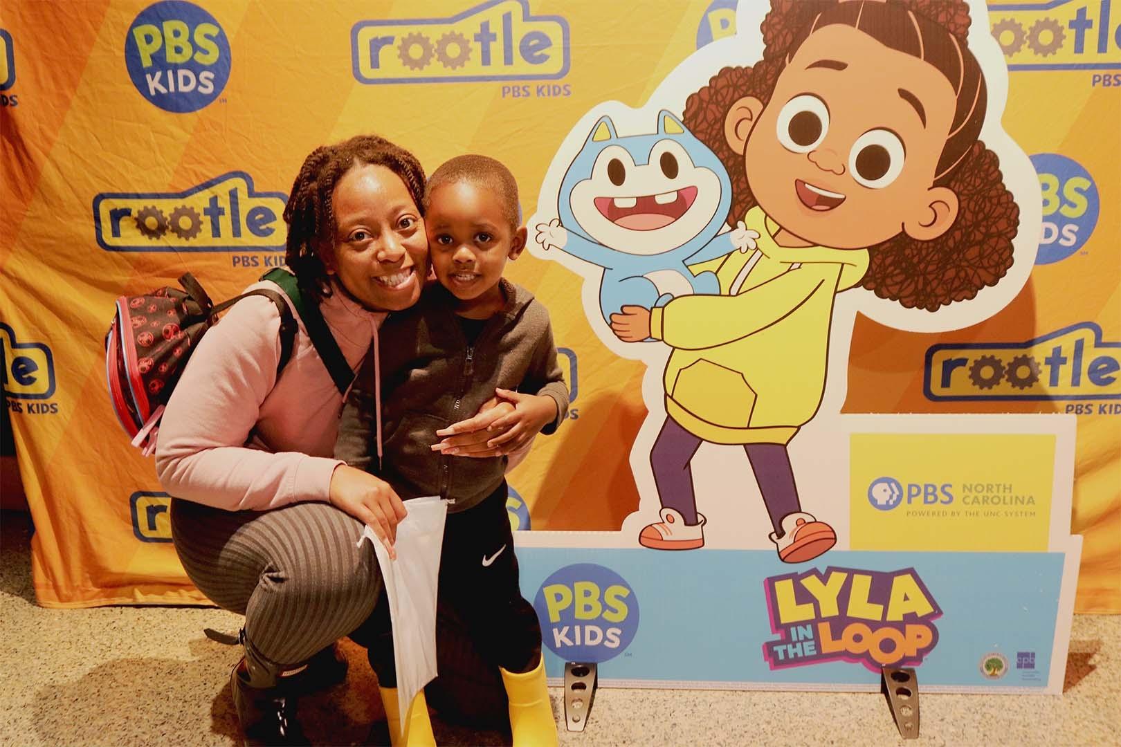 A young child and their parent pose together next to a cardboard cutout of Lyla and Stu from the PBS KIDS show, "Lyla in the Loop" at a Rootle Roadster Tour stop.