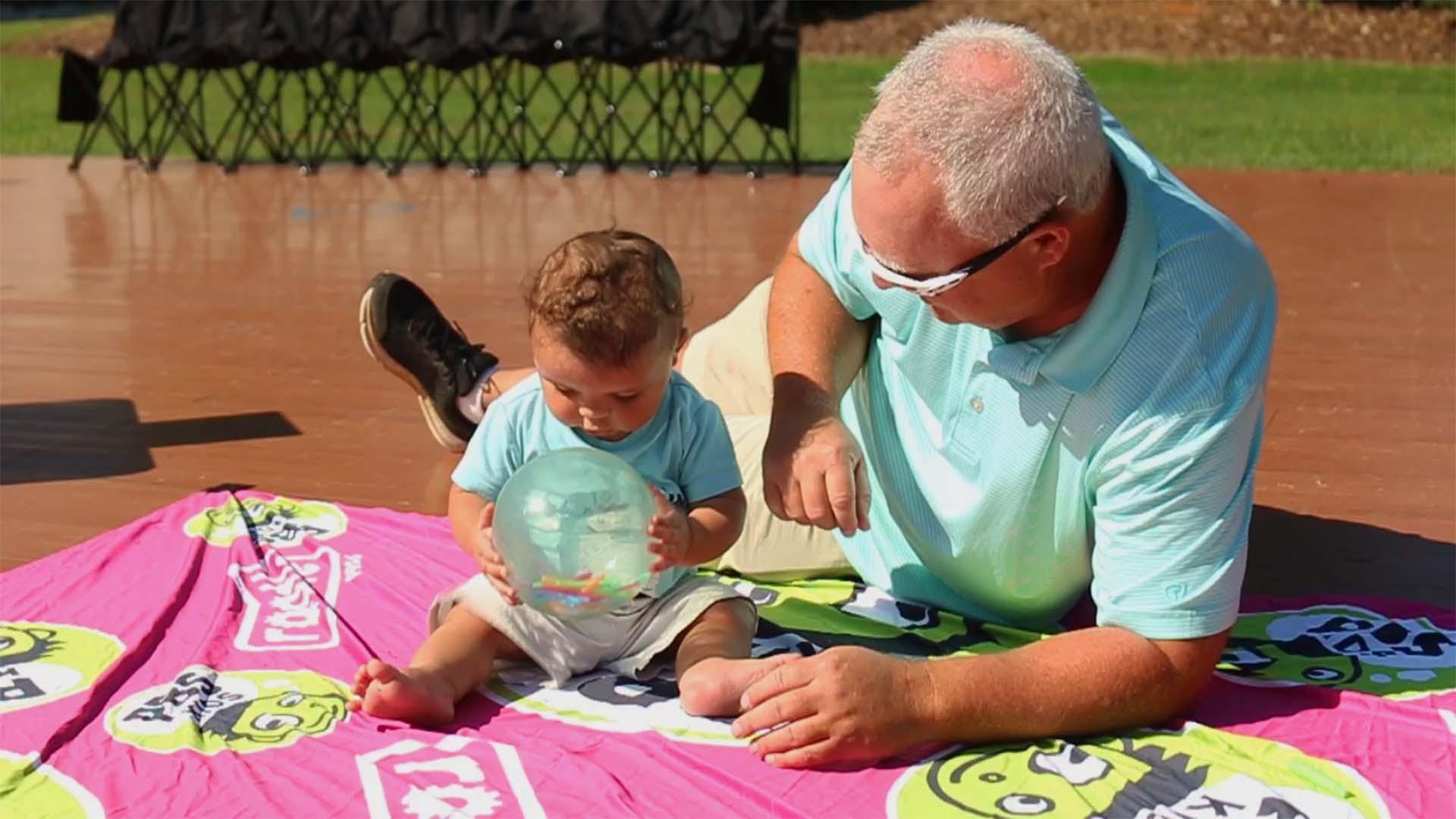 A child wearing a matching shirt with their parent as they play on a Rootle PBS KIDS picnic blanket on the ground.