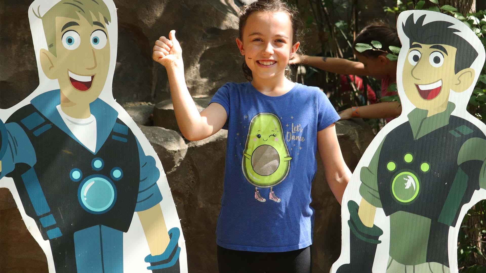 A child poses with a "thumbs up" next to cutouts of the Wild Kratt brothers.