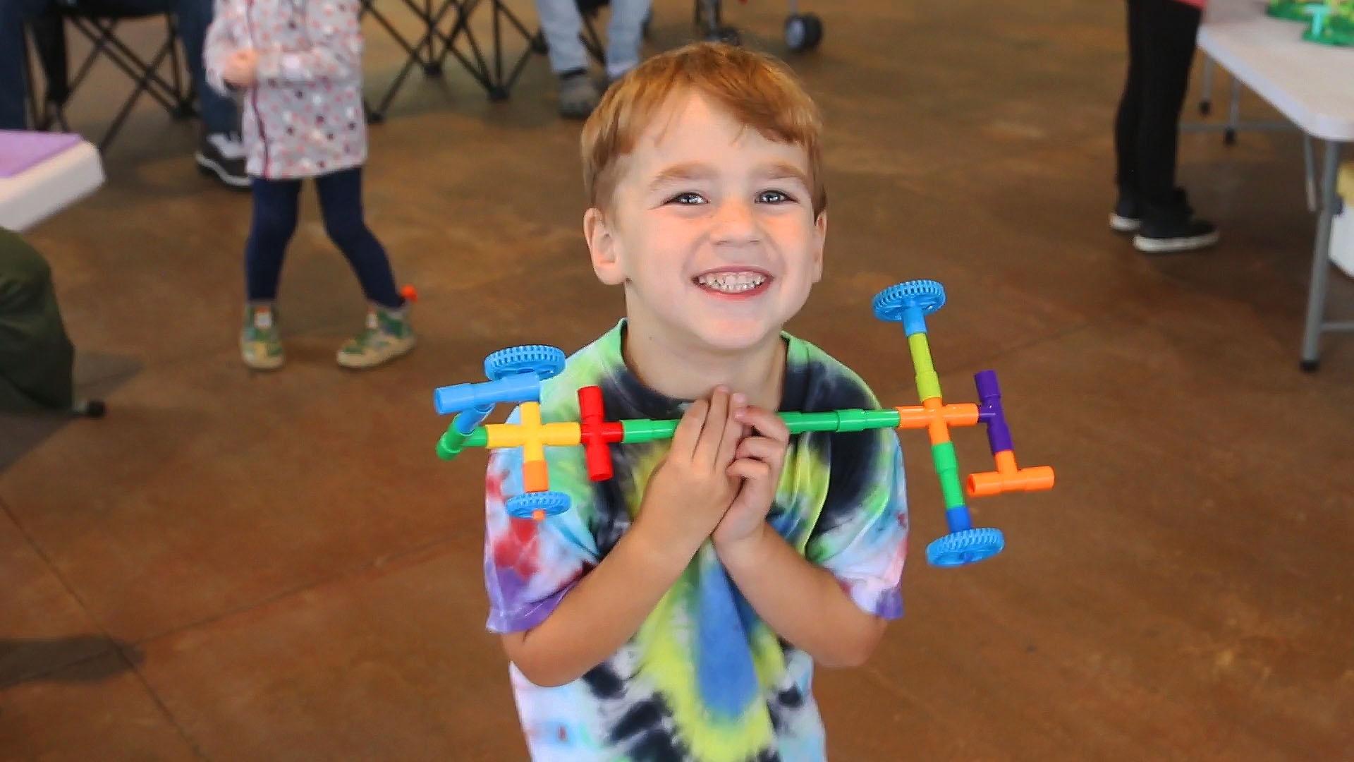 A child smiling while holding a car they made out of tubular building toys.