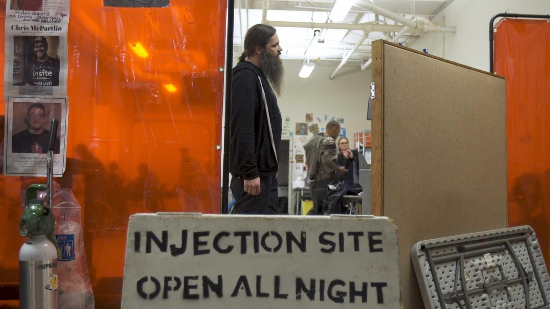 A man stands behind a sign that reads "Injection Site Open All Night"