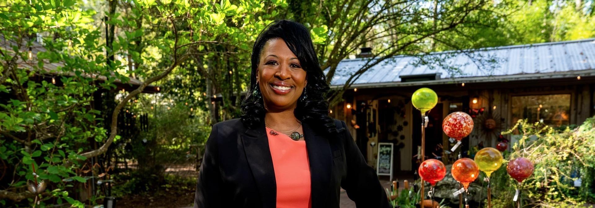 A smiling woman stands outdoors in a leafy garden setting, wearing a black blazer over a coral top. Behind her is a small shop or studio with a metal roof, string lights, and colorful glass garden ornaments displayed on poles.