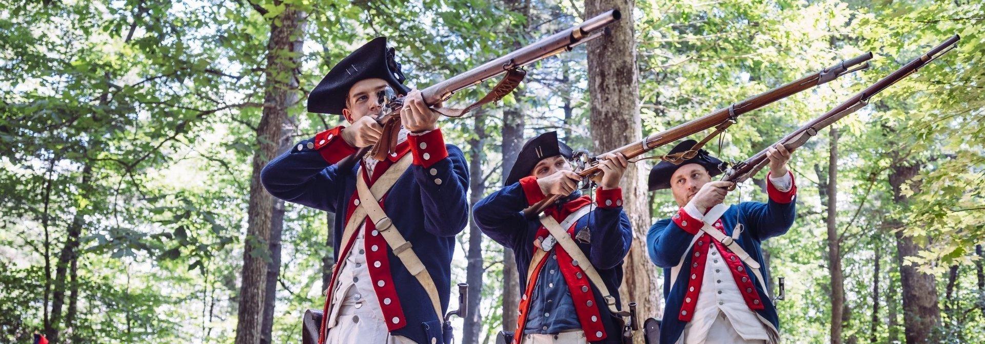 Three white men dressed in revolutionary solder uniforms and holding up rifles.