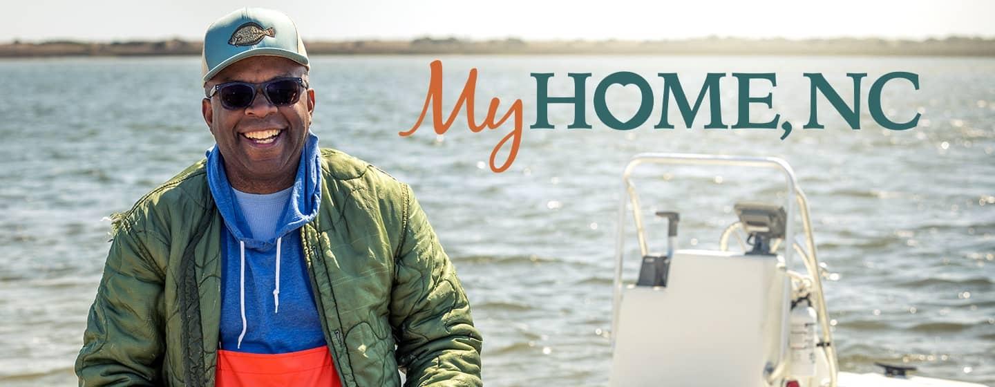 Ricky Moore in a green jacket stands on a boat on the water next to the 'My Home, NC' logo.