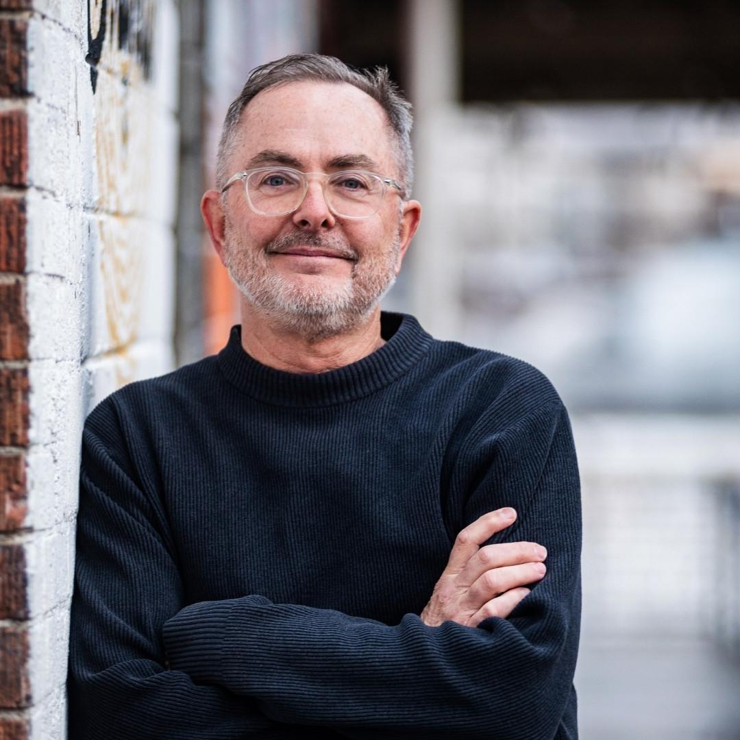 Paul Bonesteel in a black mock neck shirt leaning against a brick wall with his arms crossed.