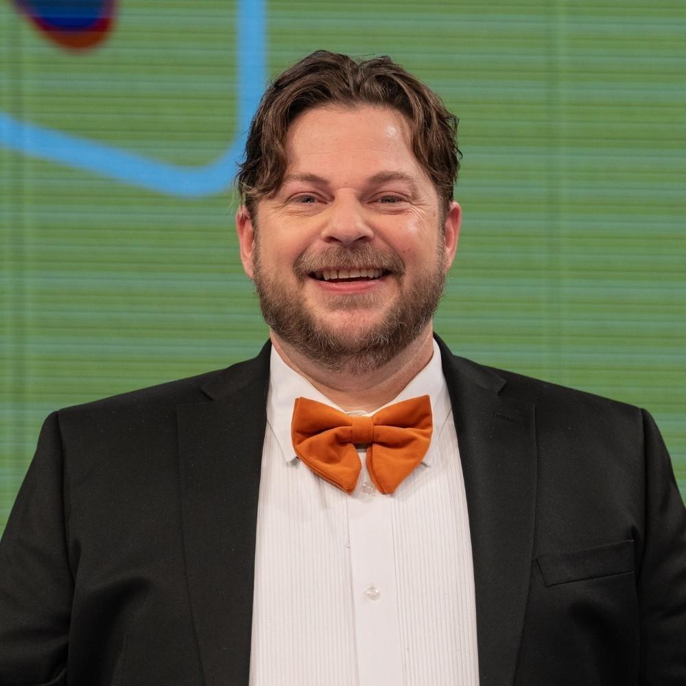 Garin Hyde, show host. A white man with a mustache and beard wearing a black suit jacket with a white button down and a large orange bowtie.