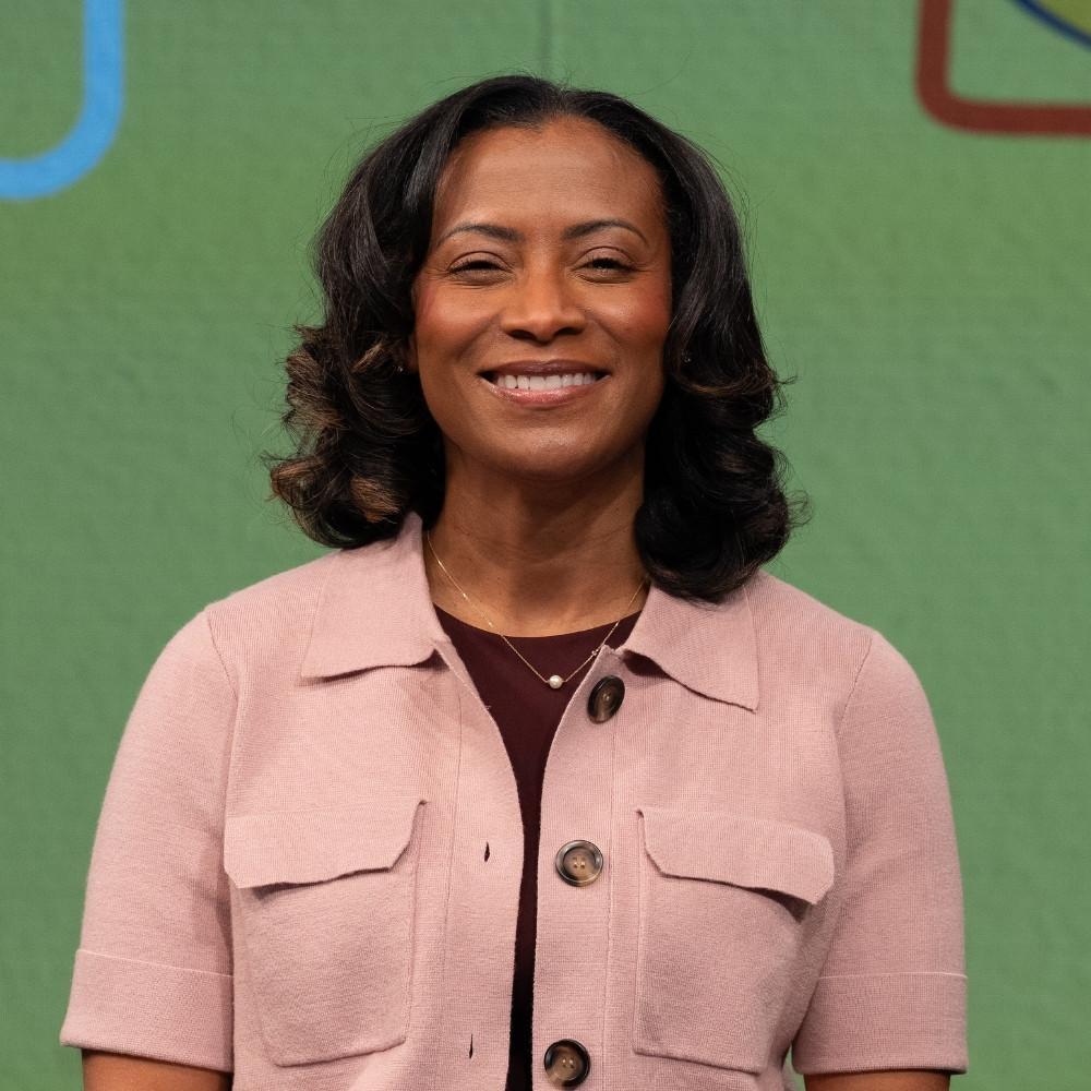 Juanella Tyler, Health Development Coordinator. A Black woman with a short, dark brown blowout, wearing a maroon shirt under a short sleeve, pink button up jacket.