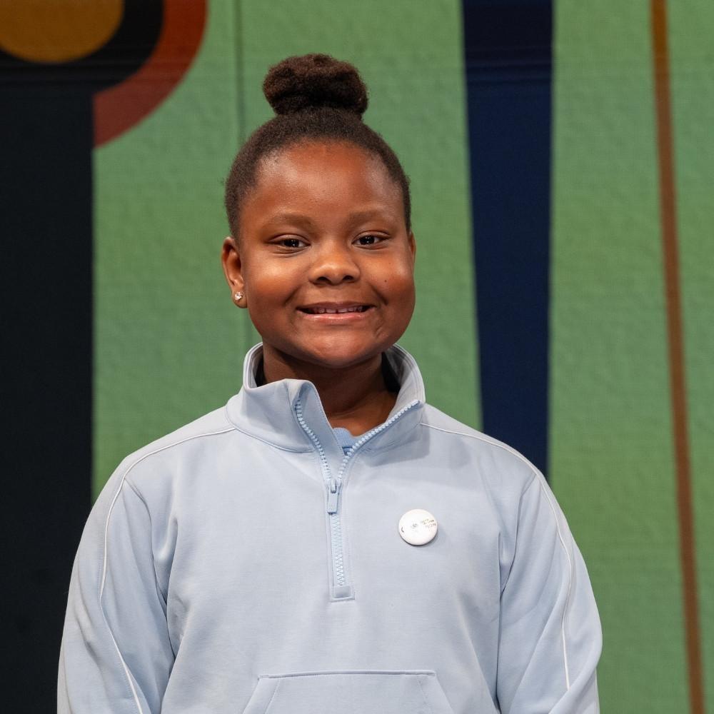 Olivia, third grader. A young Black girl wearing a grey-blue quarter zip and her hair in a tight bun.