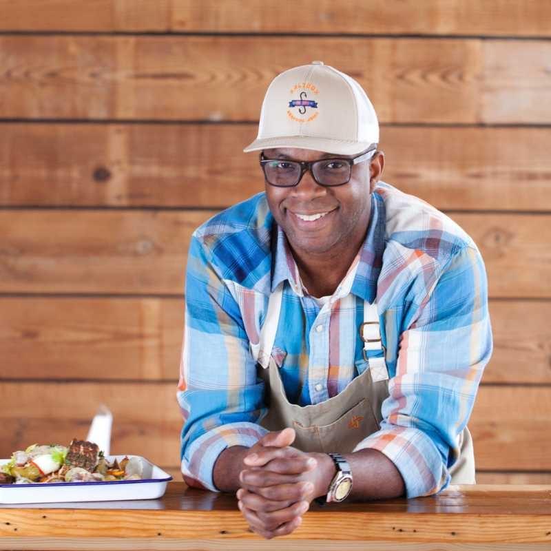 Chef Ricky Moore, wearing a bright blue plaid shirt and a Saltbox Seafood Joint baseball cap, casually leans on a table next to a dish from his restaurant