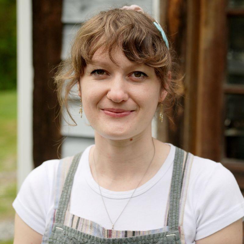 Cheryl Day smiling in front of her house's front porch while holding a white cake with a big pink peony on top.