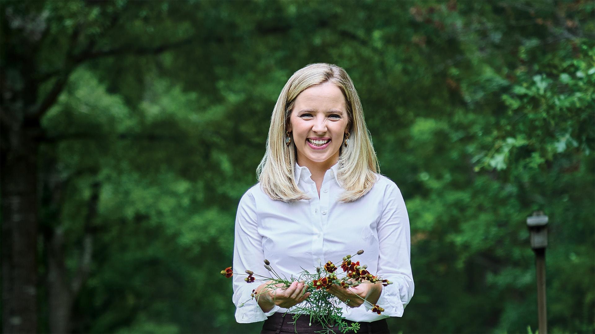 Sci NC host Laura Smith holding flowers with green trees in the background.