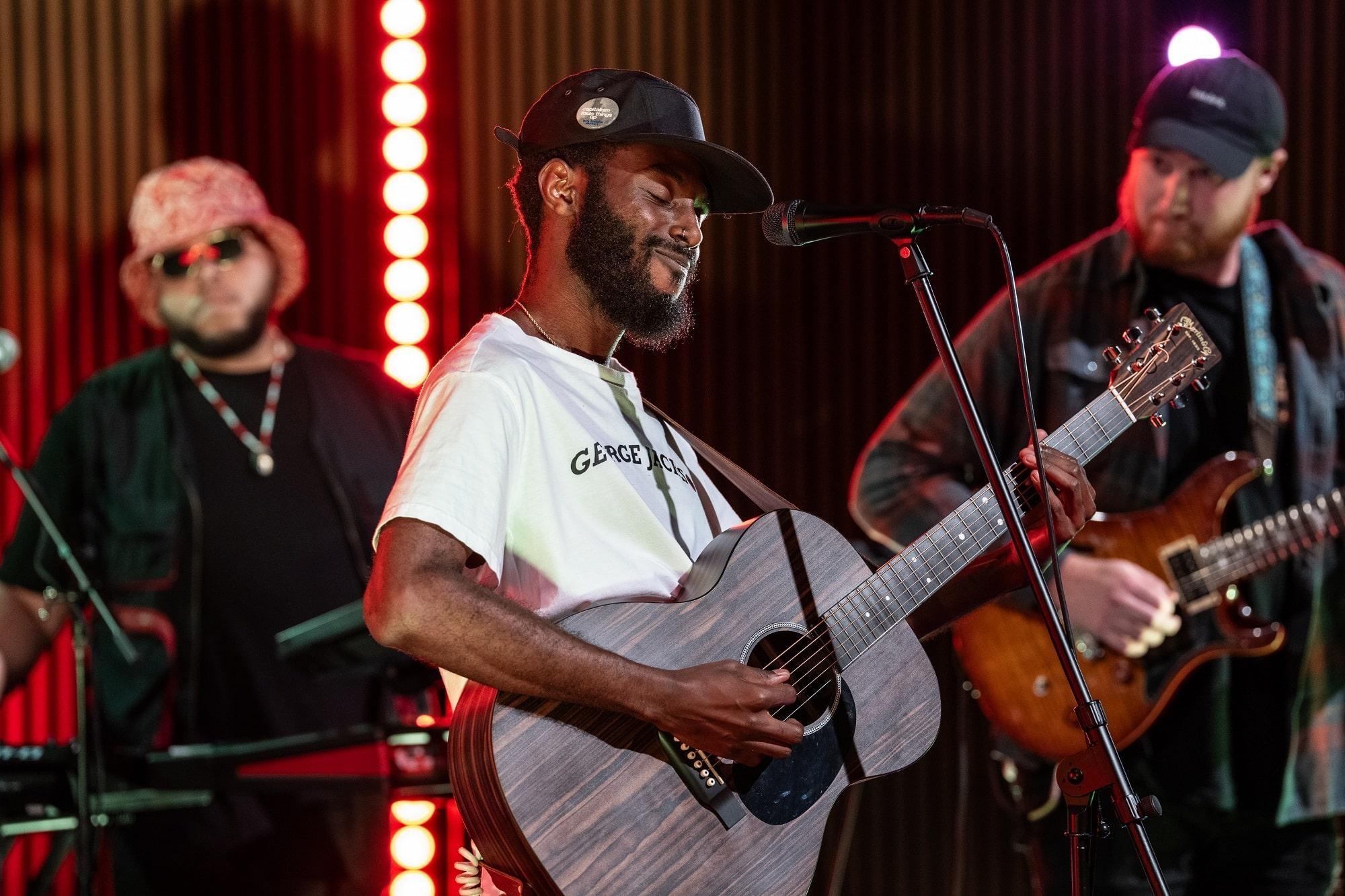 Sonny Miles plays acoustic guitar with bandmates in the background.