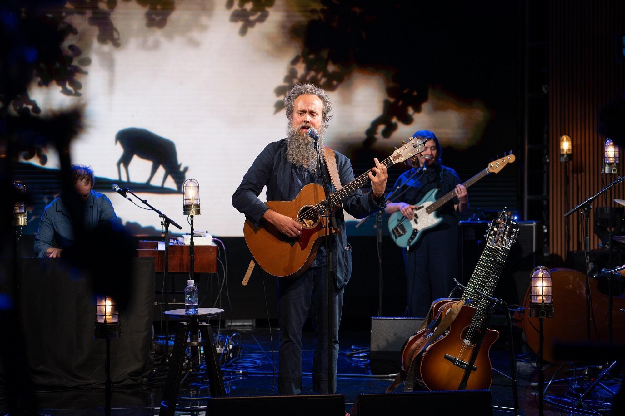 Sam Beam of Iron & Wine plays acoustic guitar while singing into a microphone.