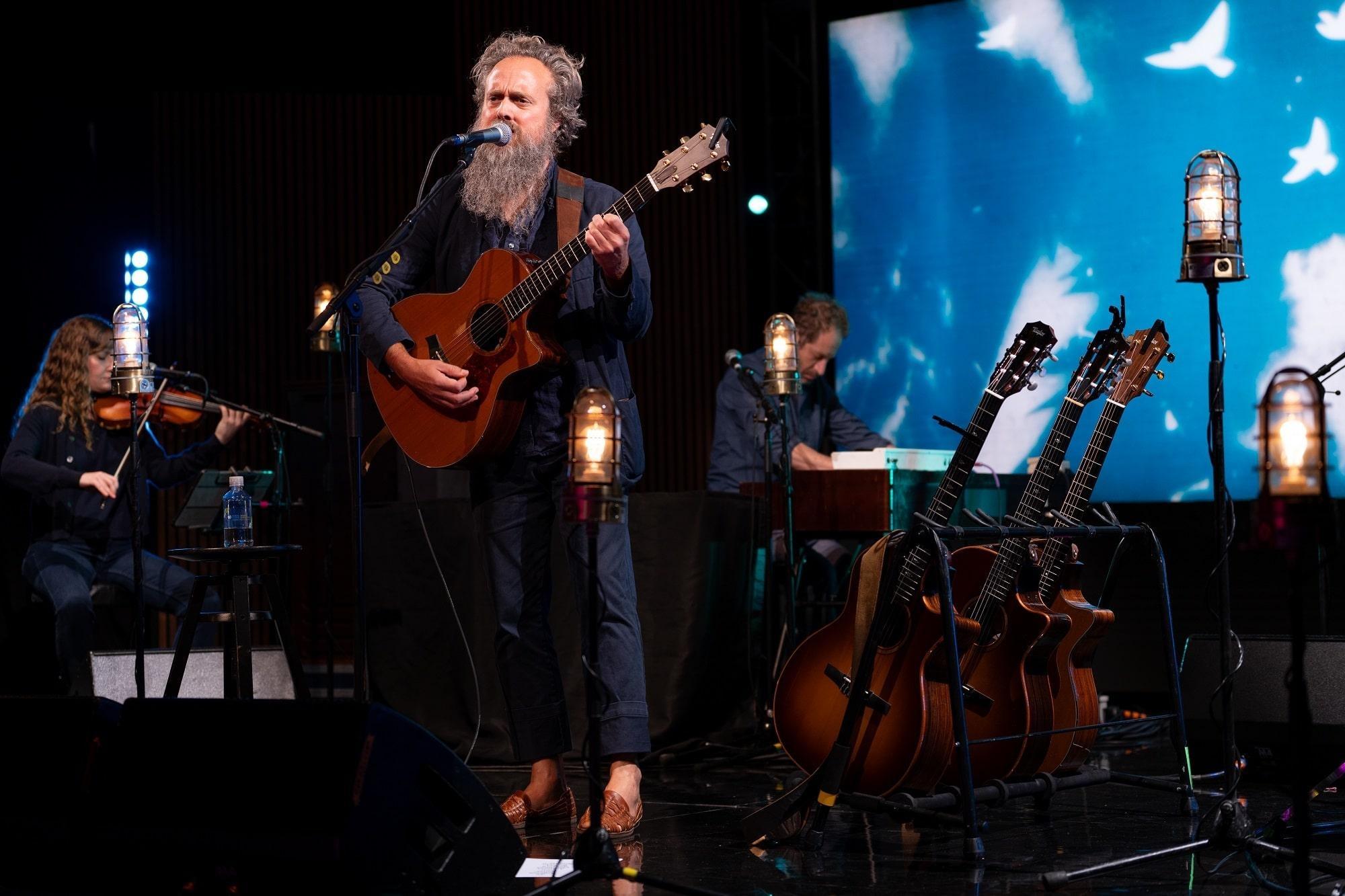 Sam Beam of Iron & Wine plays acoustic guitar while singing into a microphone.