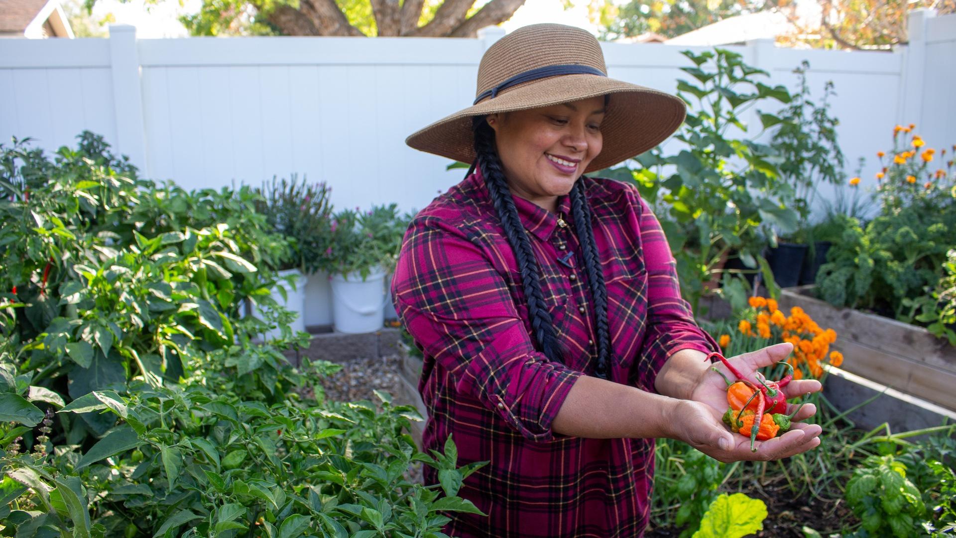 woman standing in garden with hat, braids, and plaid red shirt holding a handful of peppers
