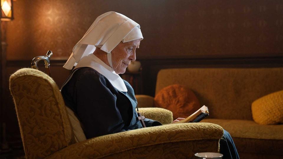 Sister Monica reads a book while sitting in a chair.
