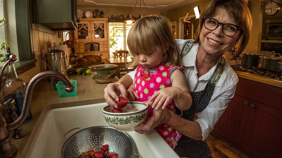 A woman holds her granddaughter up to the kitchen sink and puts washed strawberries into a bowl.