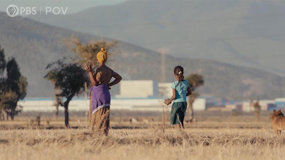 A woman and a girl stand in a field, looking at a building and mountains in the distance.
