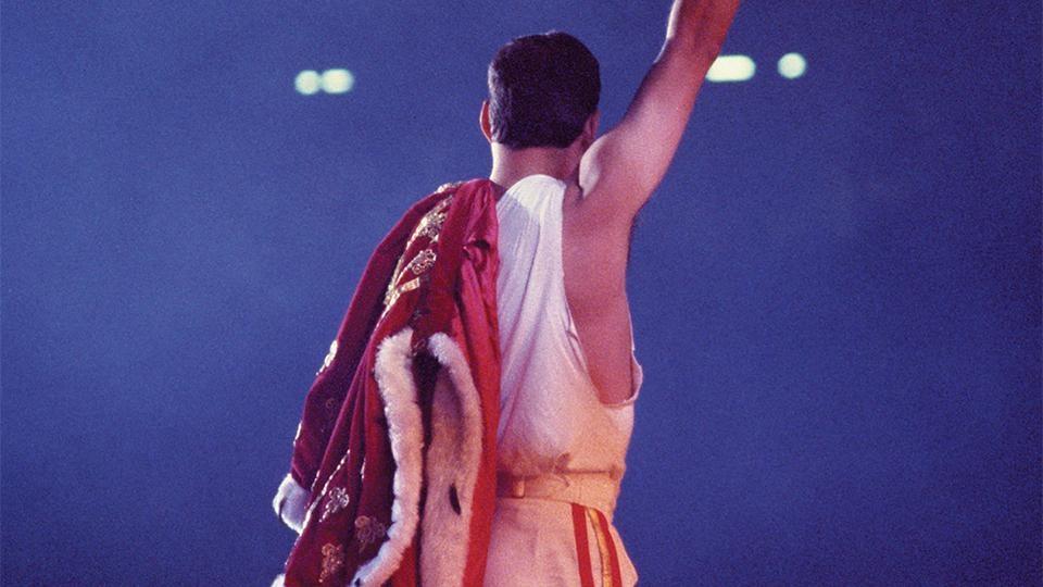 Freddie Mercury stands onstage with his back to the camera. A royal robe is slung over his shoulder.