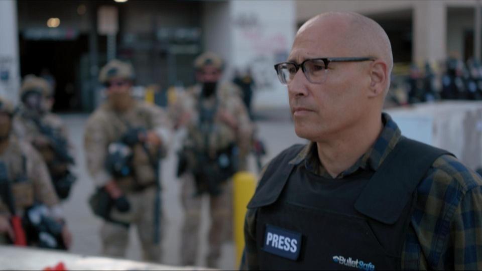 A reporter wearing a Kevlar vest stands in front of people in military garb.