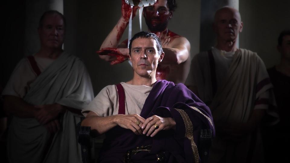 A man covered in blood stands behind Caesar.