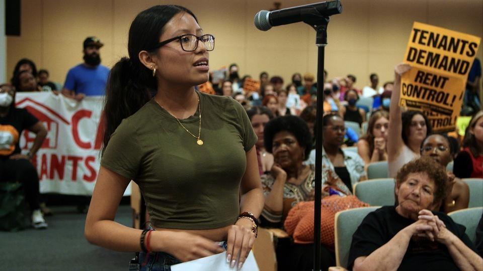 A woman stands at a microphone at a townhall meeting as people seated behind here hold up signs saying, 'Tenants Demand Rent Control.'
